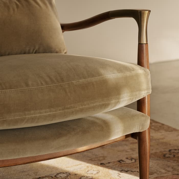 A beige, cushioned wooden armchair with brass accents is situated in a softly lit room, standing on a patterned rug. A blurred abstract wall art provides a minimalist backdrop.