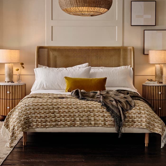 A neatly made bed with a woven headboard, checkered blanket, and pillows sits between two bedside tables holding lamps; wicker lampshade overhead; framed artwork decorates the beige wall.
