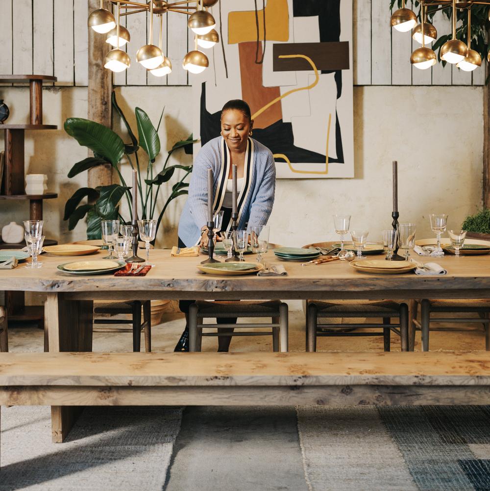 A person arranges tableware on a large wooden dining table under hanging brass lights, in a room with potted plants, abstract wall art, and rustic wooden paneling.