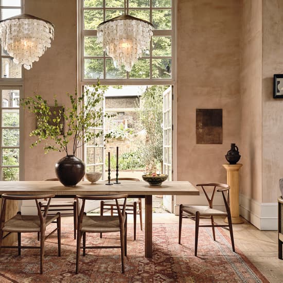 Large dining table in an elegant, sunlit room adorned with two chandeliers, surrounded by wooden chairs on a patterned rug. A vase with green branches sits on the table; French doors open to a garden.
