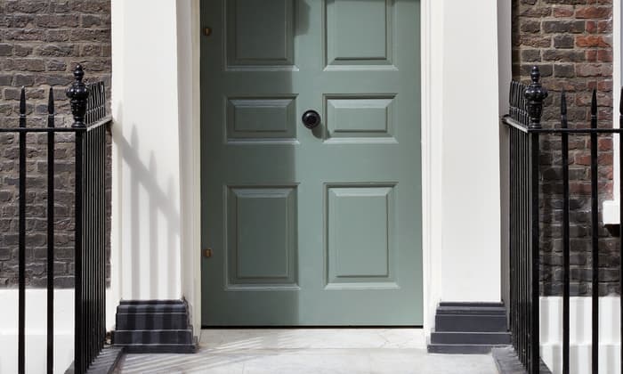 A green door labeled "76" with ornate white trim stands closed within a brick building, flanked by a black wrought-iron fence on a stone path.