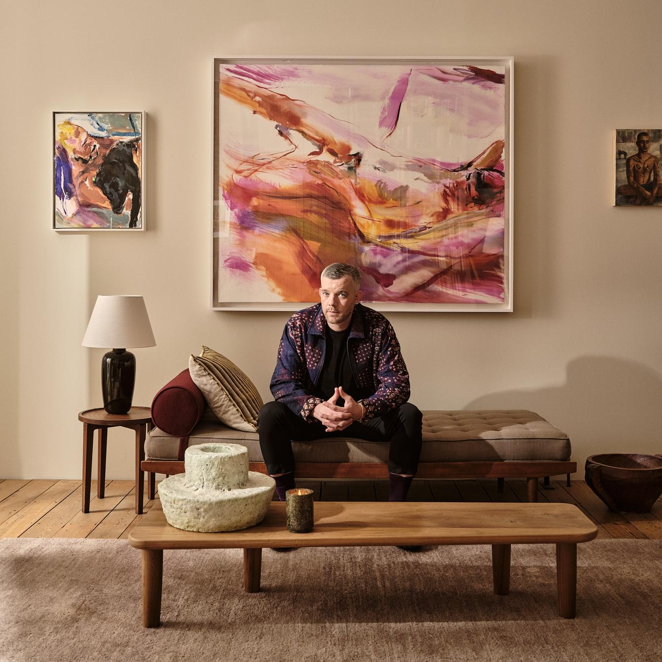 A man sits on a bench in a living room, surrounded by abstract art on beige walls, with a wooden coffee table and decor items in the foreground.