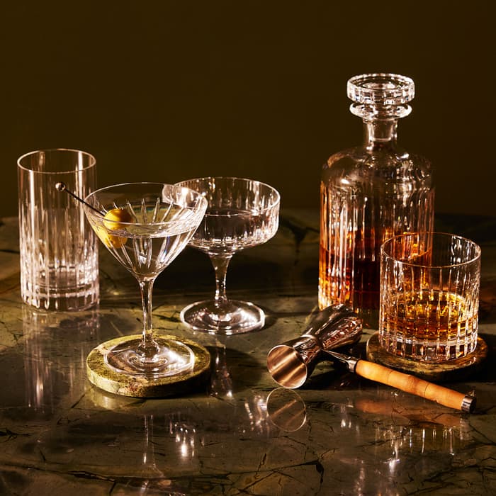 Crystal glassware filled with drinks, including a martini garnished with olives, and an ornate decanter sit on a polished marble surface, accompanied by a jigger and a bar spoon.