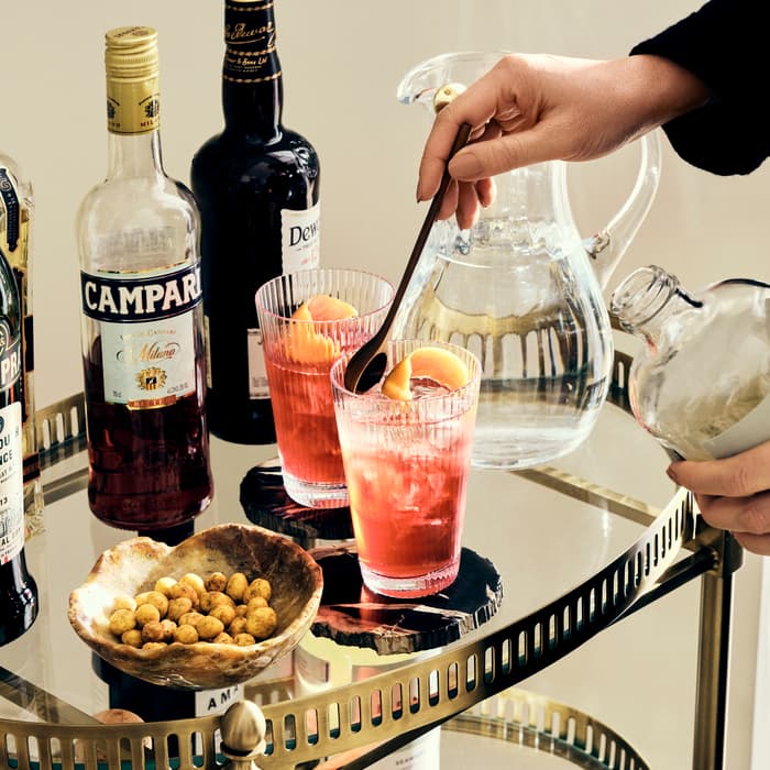Hand pours syrup into a pink cocktail garnished with an orange peel on a mirrored bar cart with various bottles of alcohol and a bowl of nuts.