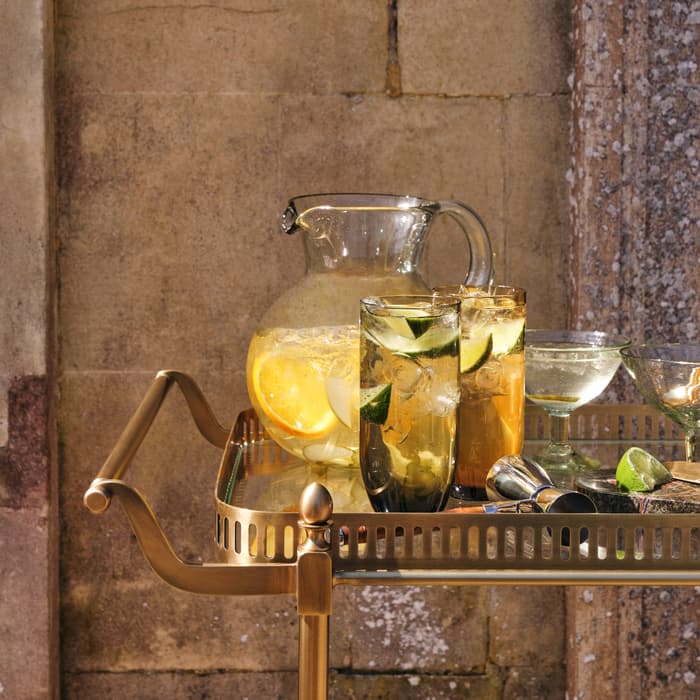 Glass pitcher and highball glasses filled with citrus-infused drinks resting on a gold trolley bar. The trolley bar is positioned against a textured stone wall bathed in natural sunlight.