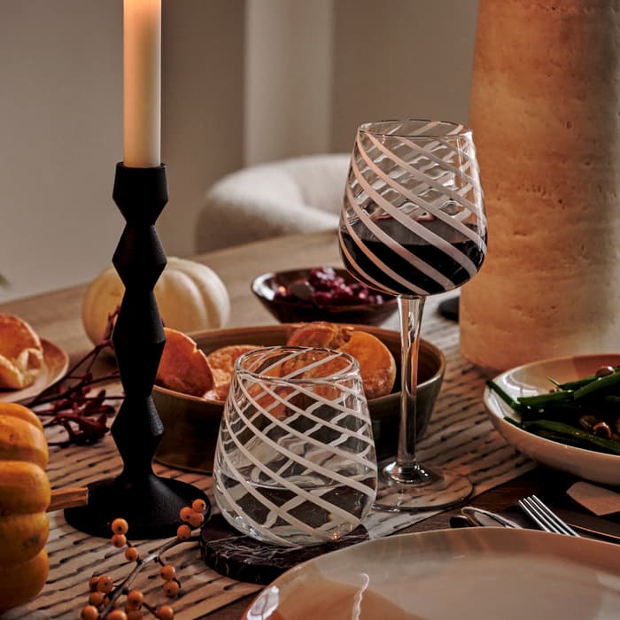 A decorated dining table featuring lit candles, patterned wine glasses with a red beverage, plates of food including green beans, rolls, and centerpiece gourds in a cozy, dimly-lit room.