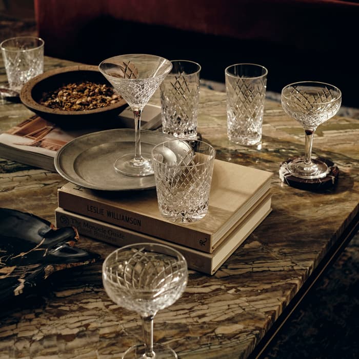 Crystal glassware, including wine and cocktail glasses, are placed alongside books and a snack bowl on a marble coffee table in a dimly lit living room with a red velvet sofa. Book titles: "LESLIE WILLIAMSON" and "The Monocle Guide to Good Business."