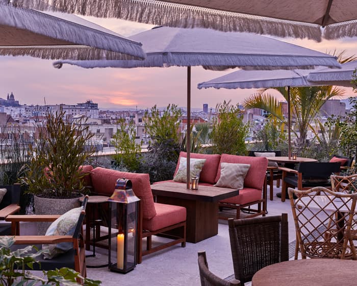 Cozy, outdoor seating with red cushioned chairs and wooden tables, shaded by large fringed umbrellas on a rooftop terrace, overlooks a cityscape at sunset, surrounded by greenery.