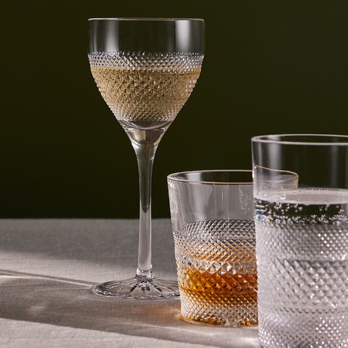 Three cut crystal glasses, one with white wine, one with amber liquid, and one with water, are placed on a linen tablecloth against a dark background.