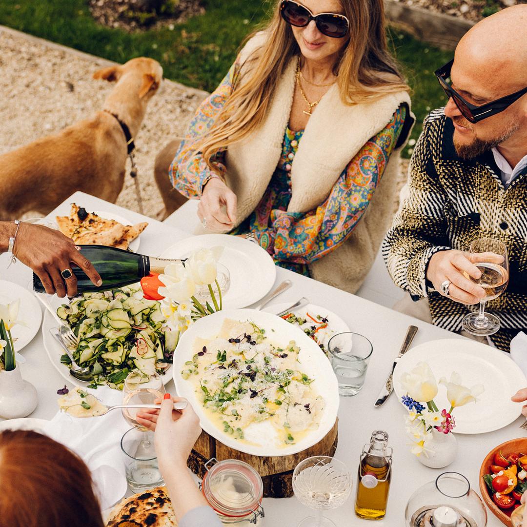 Several people dine outdoors, one pouring wine. The table is adorned with plates of food, flowers, and drinks. A dog stands beside them. The environment is grassy with gravel paths.