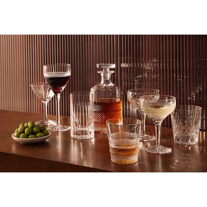 Various filled crystal glasses and a decanter are arranged on a wooden surface, accompanied by a small bowl of green olives, set against a vertically striped, translucent background.