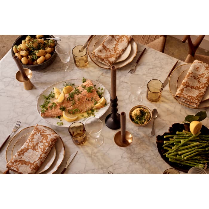 A dining table set for four features a dish of cooked salmon garnished with lemon and herbs, accompanied by boiled potatoes, asparagus, and candles on a marble surface.