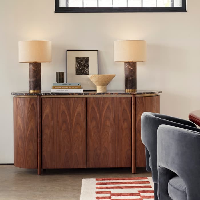 A wooden cabinet with twin lamps, books, and decor is set against a wall beneath a large arched window. Nearby, blue chairs surround a wooden table. Abstract art decorates the adjacent wall.