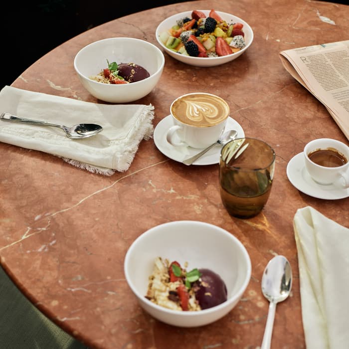 Two bowls of fruit and granola, two cups of coffee, a glass of water, and a newspaper rest on an outdoor marble table, alongside napkins and spoons.