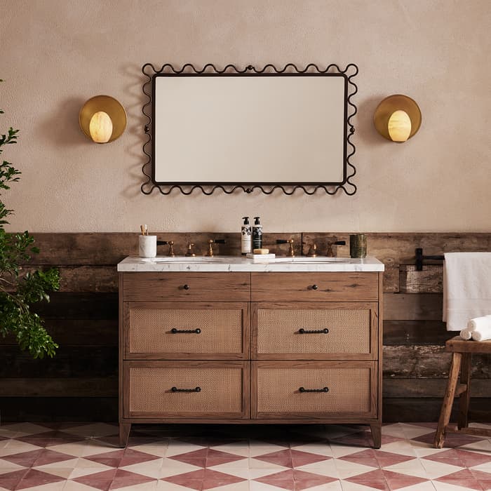 Wooden bathroom vanity with marble countertop has two circular gold wall lights and wavy-framed mirror above, beside a potted plant on a geometric-tiled floor, in a rustic setting.