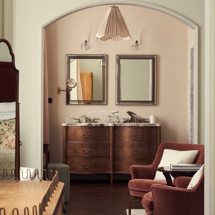Vanity with a double sink and mirrors in a cozy bathroom alcove, adjacent to a living area with plush armchairs and soft lighting.