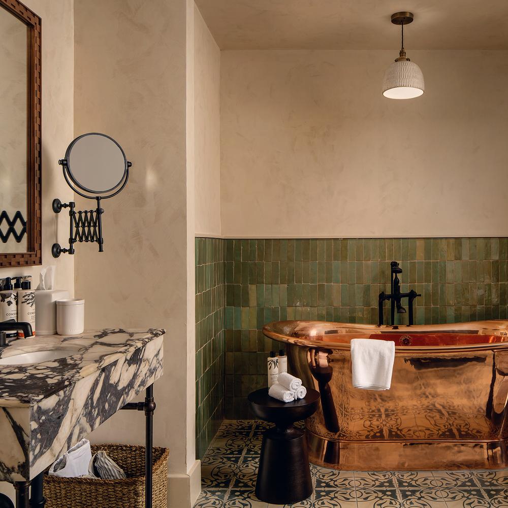 A copper bathtub, adorned with a white towel, rests in a bathroom with green-tiled walls and a patterned floor. Nearby, a marble sink counter with toiletries and a mirror is visible.