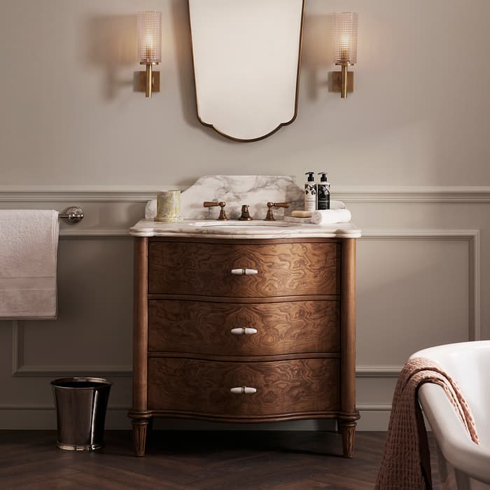 A wooden vanity with a marble countertop holds a mirror and two soap dispensers. Above, two wall sconces emit light in a bathroom featuring elegant paneling and herringbone flooring.