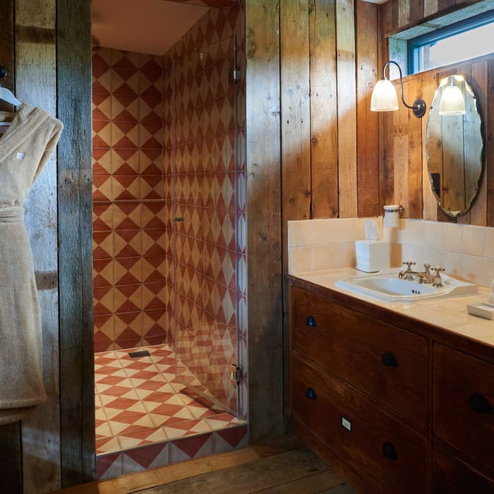 A bathroom features dual sinks on a wood vanity with a large mirror, adjacent to a shower with geometric red and white tiles, and a tan bathrobe hanging on a wooden wall.