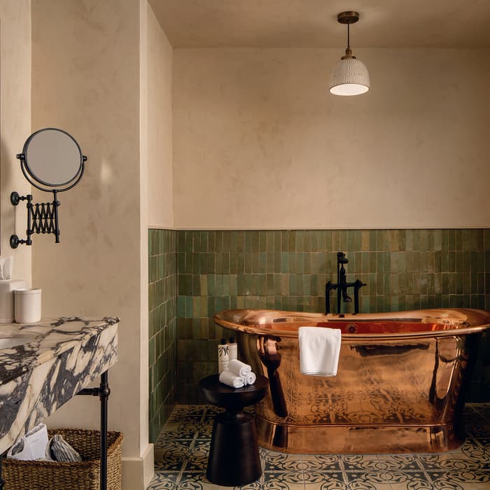 Copper bathtub with a towel hanging over it, set against green tiled walls. Marble sink with products, mirror, and magnifying mirror. Black stool with rolled towels and a plant.
