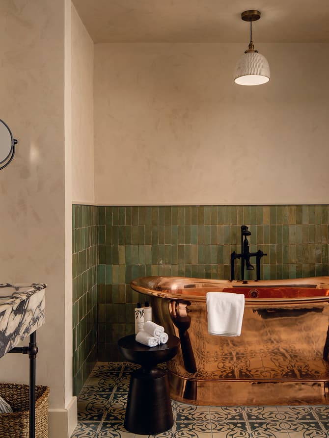 Copper bathtub with a towel hanging over it, set against green tiled walls. Marble sink with products, mirror, and magnifying mirror. Black stool with rolled towels and a plant.
