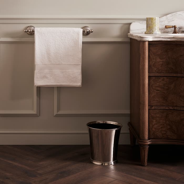White hand towel hanging on a silver rod, next to a wooden cabinet with a marble top, in a bathroom with beige walls and herringbone wood flooring. Silver trash bin below.