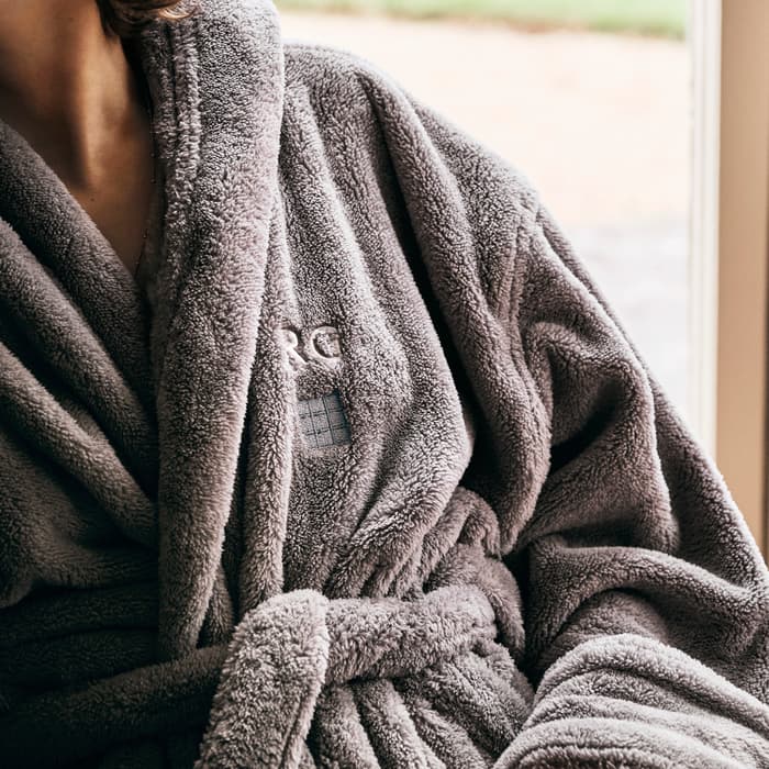 A person wearing a gray plush robe sits indoors, holding a brown mug with a hot beverage. The robe has embroidered text "RC" on it. The outside view shows greenery.