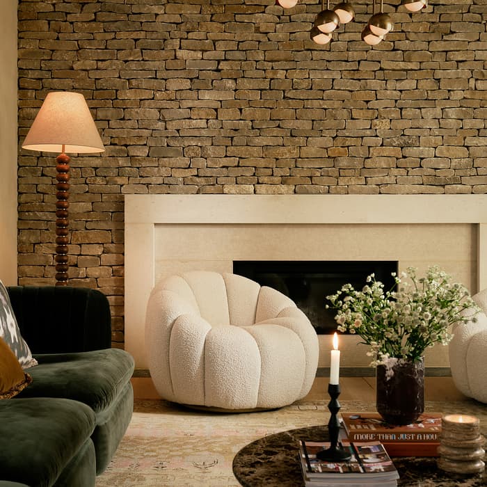 A cozy living room features a stone fireplace, two rounded white chairs, a dark green sofa, a floor lamp, and a round table with flowers, a lit candle, and stacked books.