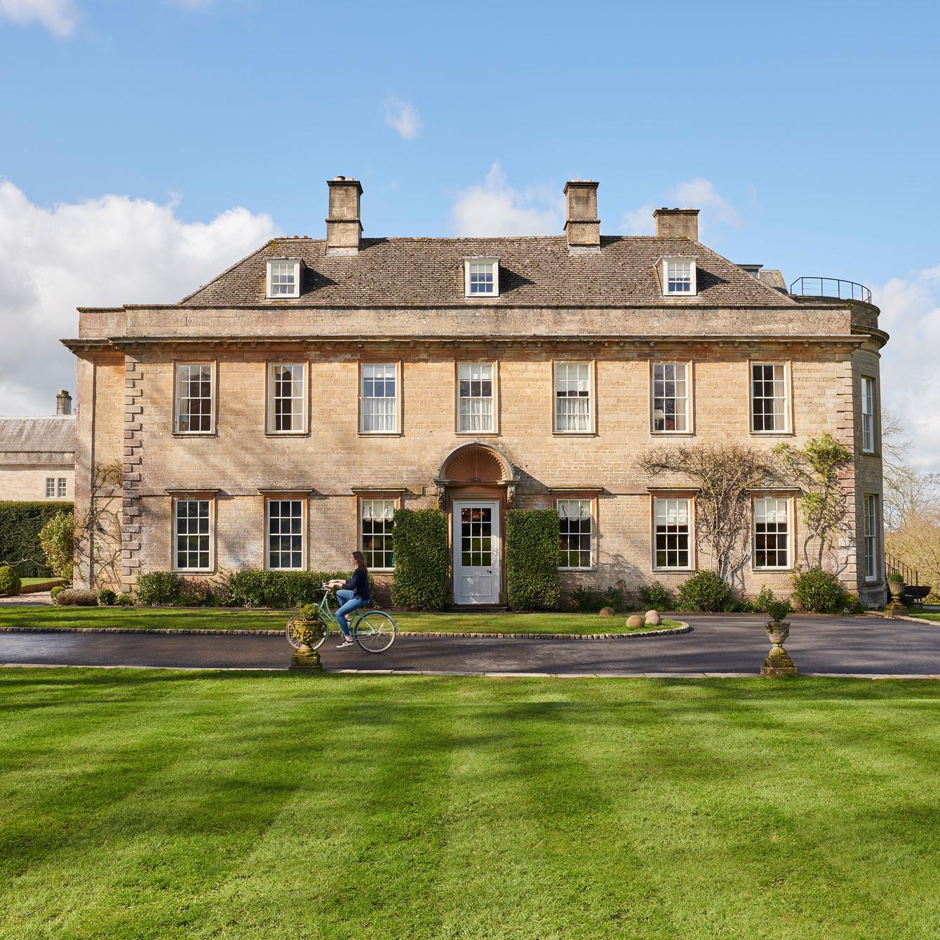 A person rides a bicycle past a large, symmetrical, three-story stone mansion with multiple windows in a landscaped garden with neatly trimmed grass and hedges under a bright blue sky.
