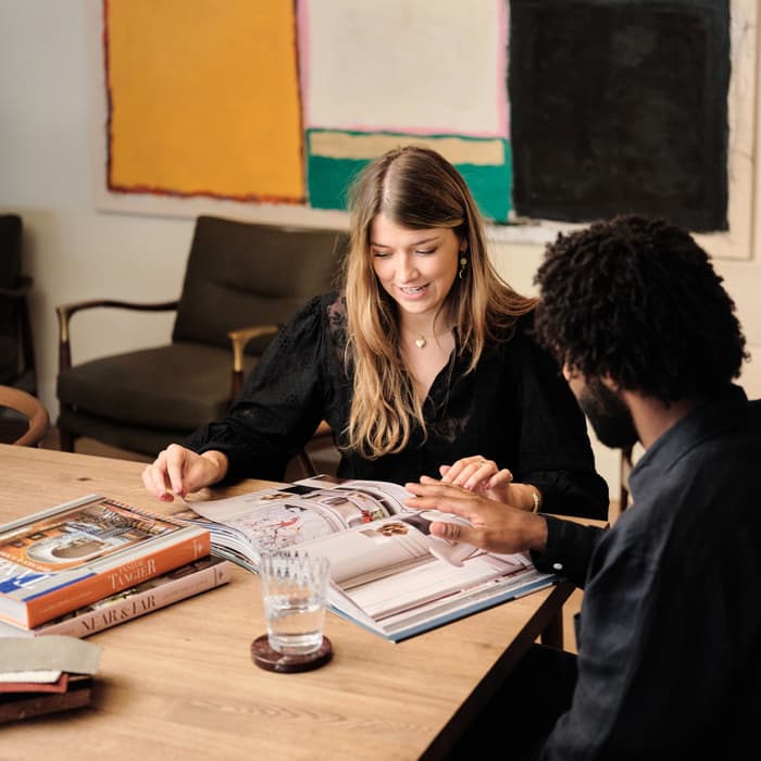 Two people sit at a wooden table, looking at a book with several other books, a water glass, and coasters. A colorful abstract painting hangs on the wall behind them.