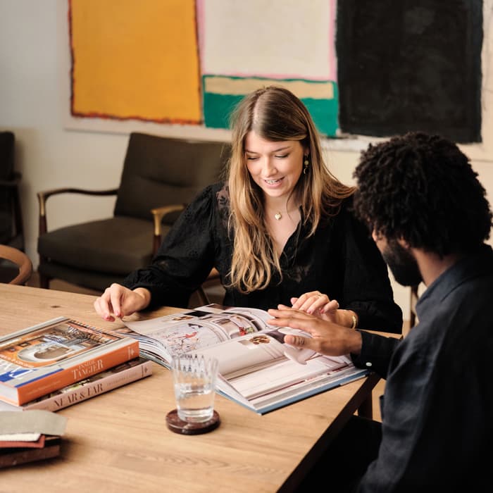 A woman and a man examine a large open book on a wooden table, surrounded by additional books and a glass; they are seated in a room with modern abstract art on the wall.