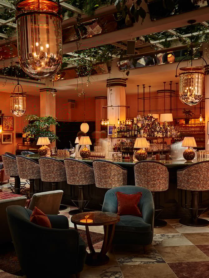 A large, elegant bar with cushioned, patterned stools, illuminated by hanging vintage lamps, surrounded by lush indoor plants, cozy armchairs, and small round tables in a warm, sophisticated setting.