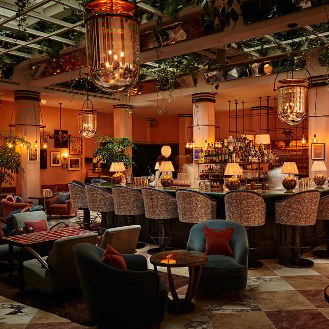 A large, elegant bar with cushioned, patterned stools, illuminated by hanging vintage lamps, surrounded by lush indoor plants, cozy armchairs, and small round tables in a warm, sophisticated setting.