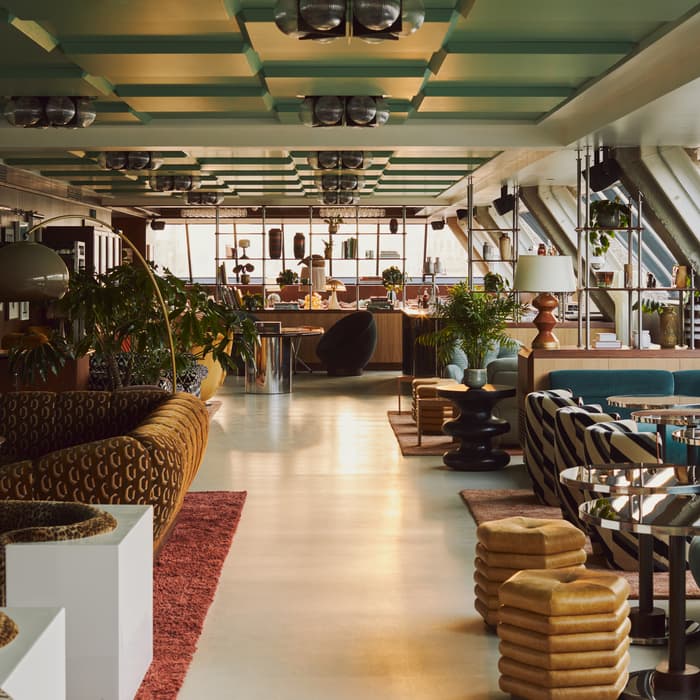 Lounge area featuring eclectic seating arrangements with patterned sofas and contemporary chairs, accompanied by stylish tables. Indoor plants and decorative objects enhance the space, illuminated by natural light through slanted windows.