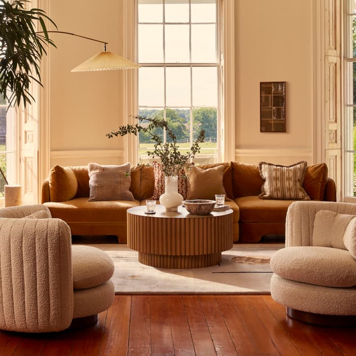 A cozy living room features a mustard-colored velvet sectional adorned with pillows, a round wooden coffee table with decor, plush armchairs, and large windows offering a scenic view outside.