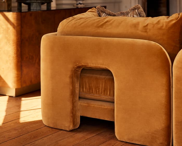 A plush, mustard-colored armchair with a rectangular cut-out in a stylish, sunlit living room, surrounded by ceramic vases, a side table with a lamp, and a potted plant.