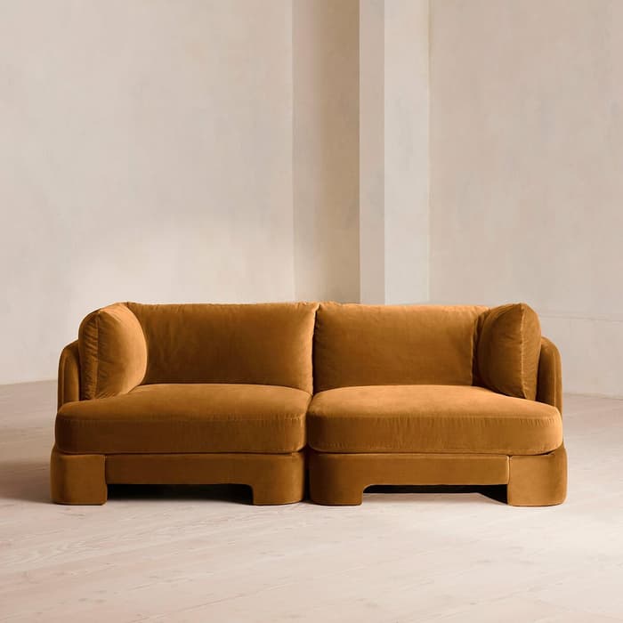 A mustard brown sectional sofa with plush cushions sits in a minimalist room with light beige walls and a light wooden floor.