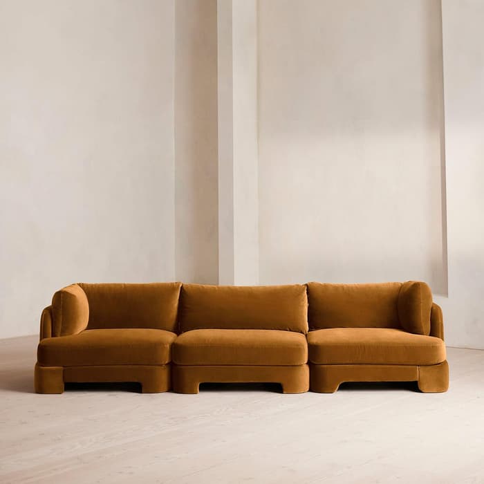 Brown sectional sofa sitting in a minimally designed, high-ceilinged, off-white room with light wooden flooring.