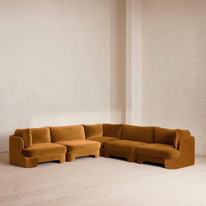 A mustard-colored sectional sofa with five plush cushions sits on a light wooden floor against a plain, cream-colored wall in a spacious, minimally decorated room.