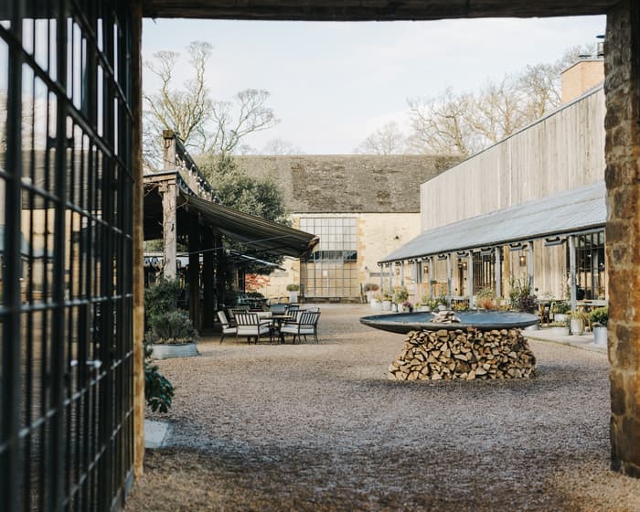 Outdoor courtyard features metal chairs around tables, surrounded by rustic brick and glass buildings with large windows, shaded awnings, and greenery, with a central stone fountain or fire pit on a woodpile.