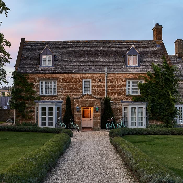 A two-story stone house with ivy on one side, lit warmly from inside, stands among manicured hedges and lawns with several bicycles near the entrance at dusk.
