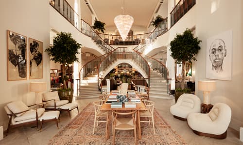 A wooden table with books and chairs sits on a rug in a luxurious room featuring a grand double staircase, large artwork, lush plants, and elegant seating.