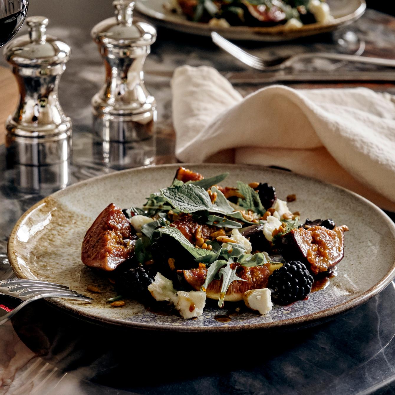 A plated salad with figs, blackberries, leafy greens, goat cheese, and nuts rests on a marble table, accompanied by a glass of red wine, cutlery, two metal shakers, and a cloth napkin.